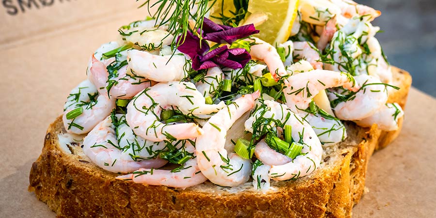 A traditional Danish open faced sandwich, knows as a Smørrebrød. It consists of a crusty slice of thick bread topped with pink prawns, green dill and garnished with a slice of lemon.